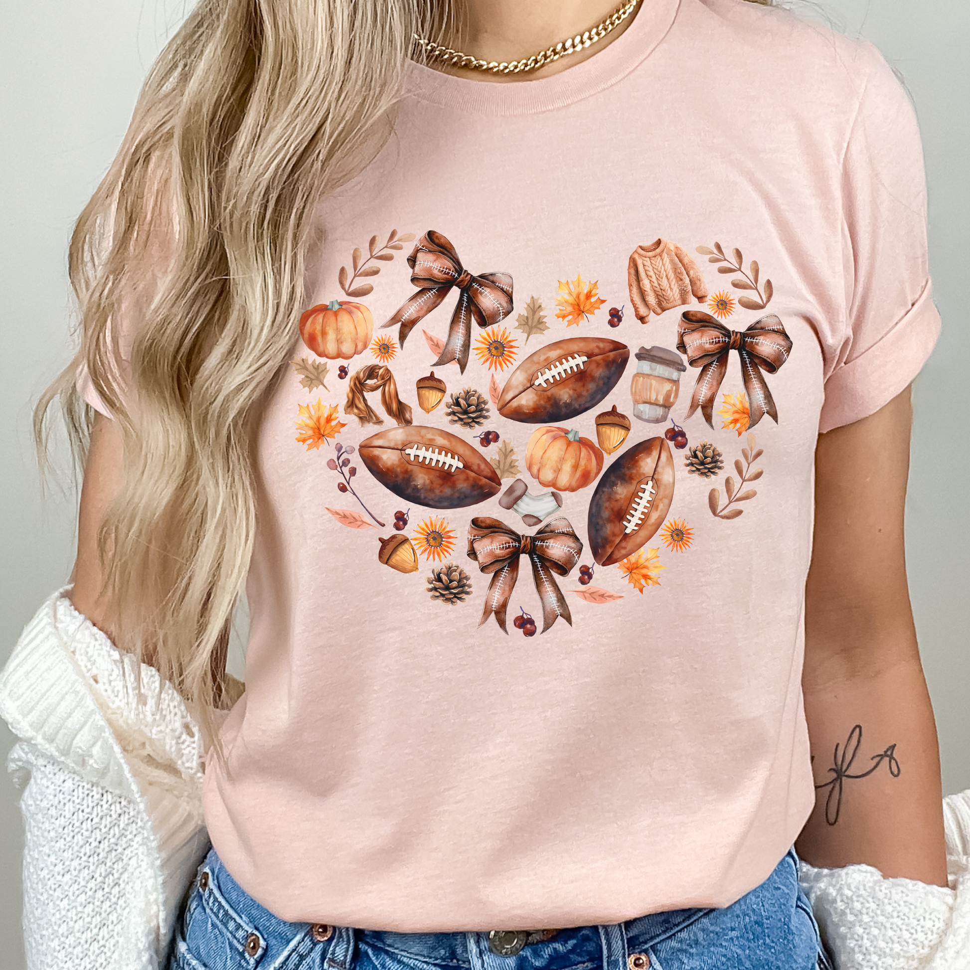 Fall Football & Pumpkin Bow Heart Unisex Shirt in pastel pink. The shirt features a beautiful autumn design with footballs, pumpkins, acorns, bows, and leaves arranged in a heart shape. Perfect cozy tee for fall, football season, and game day events.