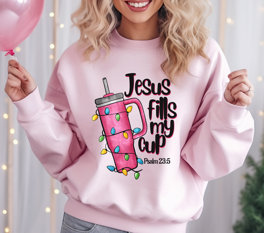 Light pink sweatshirt featuring a tumbler wrapped in Christmas lights with the text “Jesus Fills My Cup” and Psalm 23:5