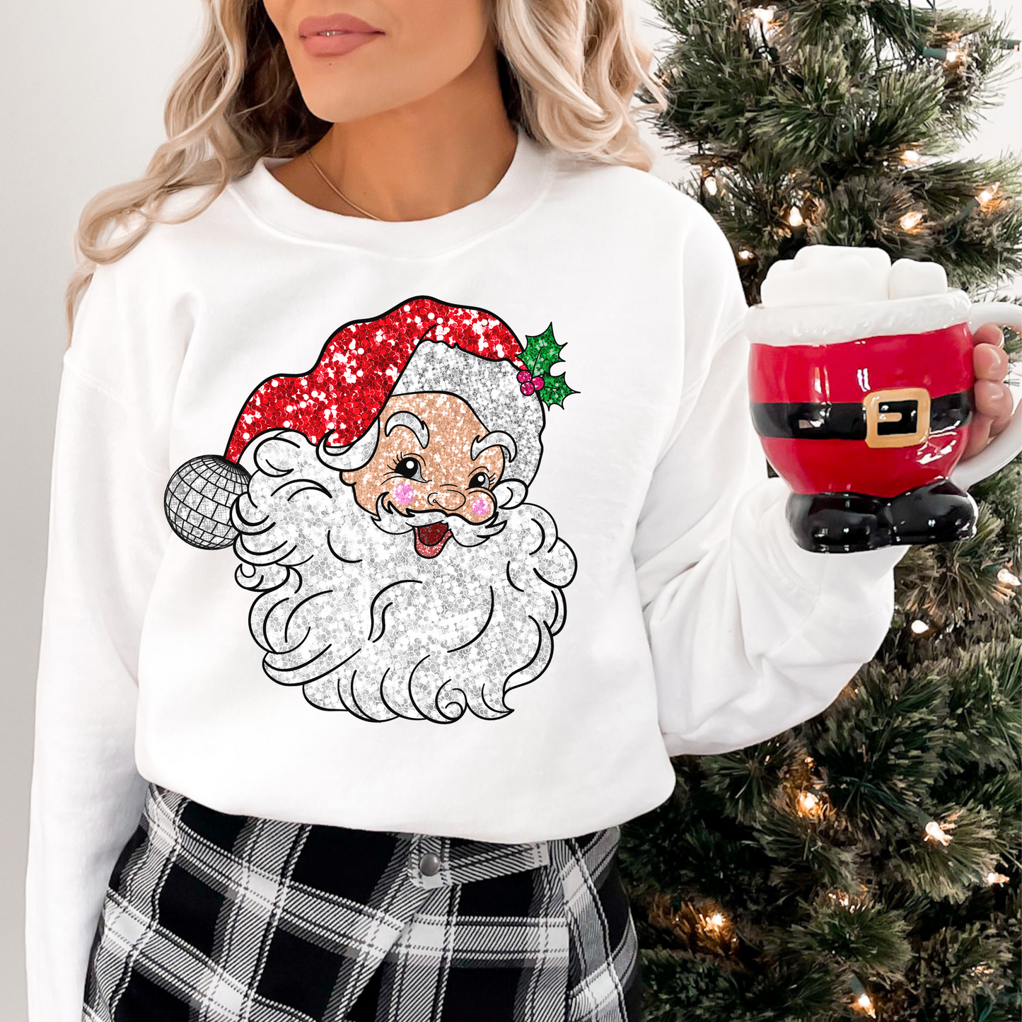 Woman wearing a white sweatshirt featuring a retro Santa Claus graphic with a sparkling red hat and disco ball ornament, holding a red Santa-shaped mug, standing next to a Christmas tree decorated with lights, showcasing a cozy and festive holiday look.