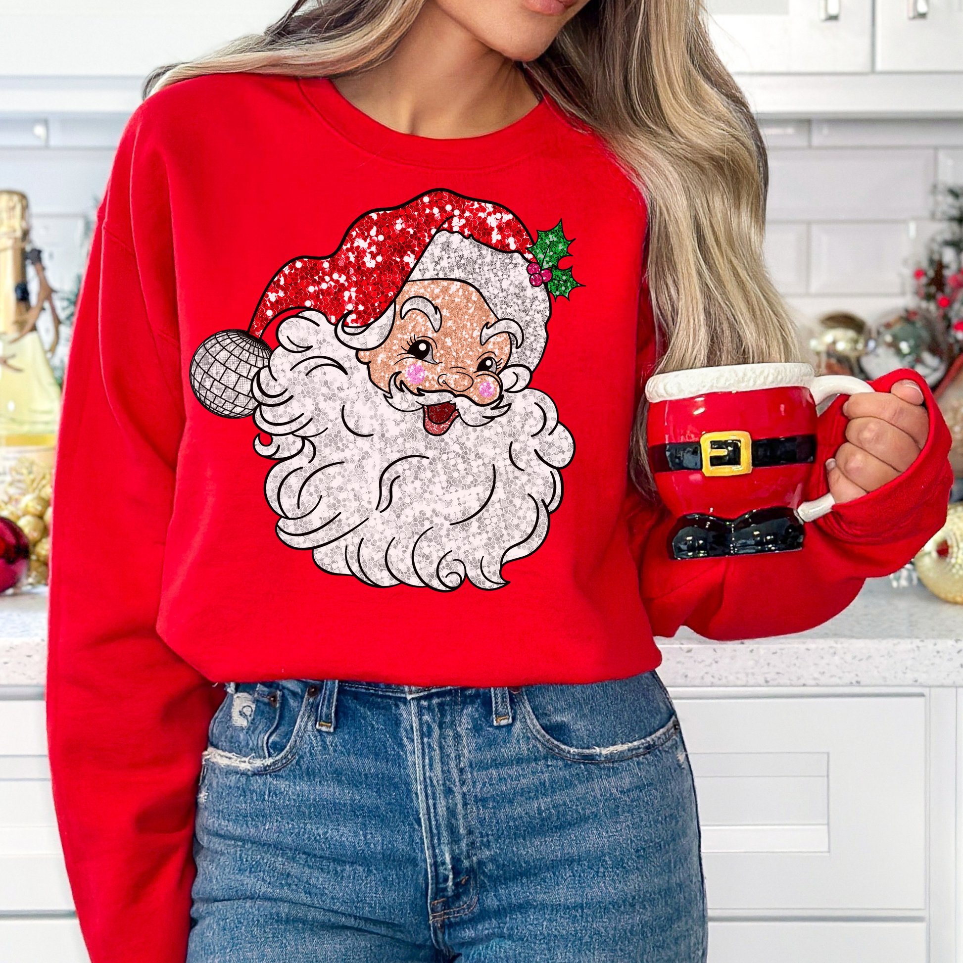 Woman wearing a white sweatshirt featuring a retro Santa Claus graphic with a sparkling red hat and disco ball ornament, holding a red Santa-shaped mug, standing next to a Christmas tree decorated with lights, showcasing a cozy and festive holiday look.