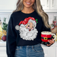 Woman wearing a white sweatshirt featuring a retro Santa Claus graphic with a sparkling red hat and disco ball ornament, holding a red Santa-shaped mug, standing next to a Christmas tree decorated with lights, showcasing a cozy and festive holiday look.